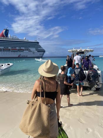 People on a sandy beach boarding a small boat, with a large cruise ship in the background, under a partly cloudy sky