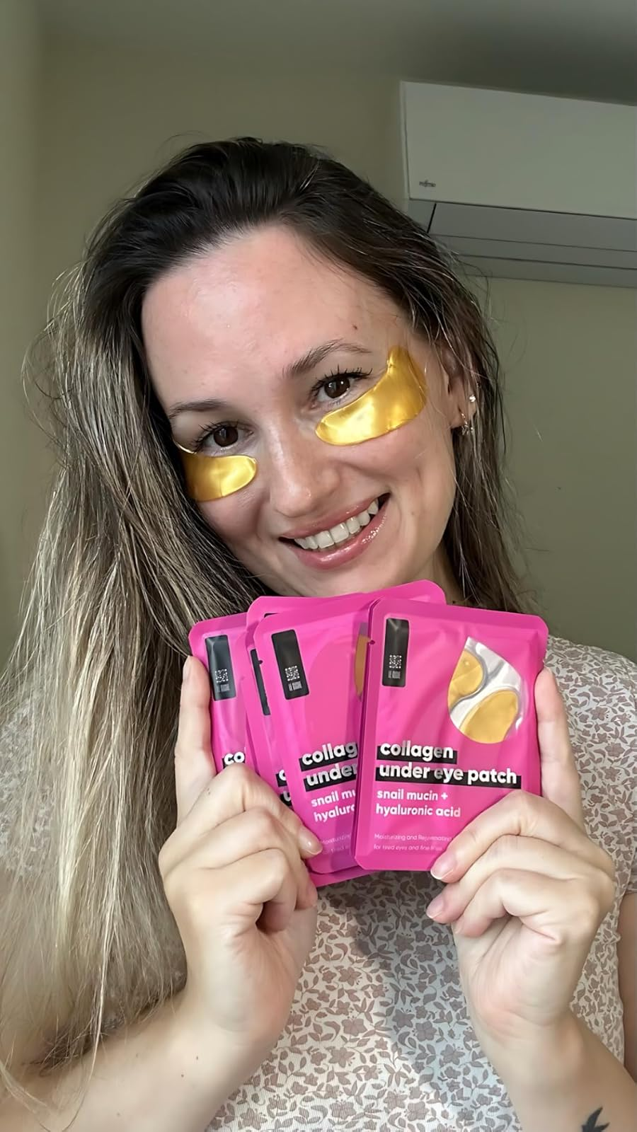 Person smiling, wearing gold under-eye patches, holding packages of collagen under-eye patches visible