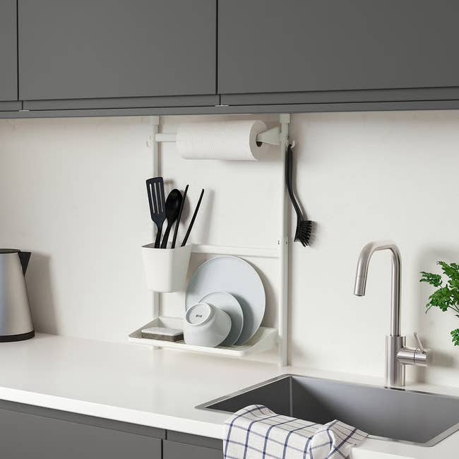 Kitchen setup with a wall-mounted organizer holding a paper towel roll, utensils, and plates above a sink, designed for efficient storage