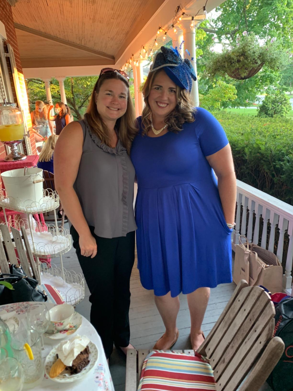 Two radical   smiling connected  a porch. One wears a ruffled apical  and pants; the different   a bluish  formal  with a matching fascinator, suitable for a societal  gathering