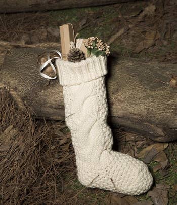 Knit stocking with small gifts and pinecone resting on logs, featured in a shopping article about holiday decor or gifts