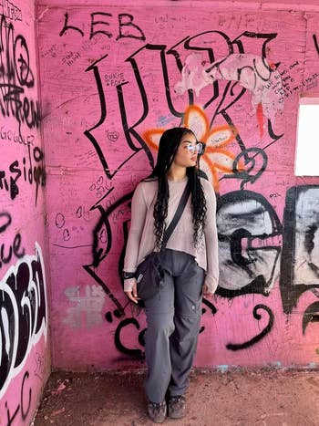 Woman with braided hair wearing glasses, a light top, and gray pants, standing against a graffiti-covered wall
