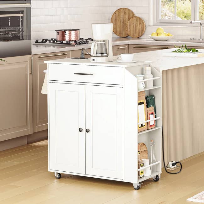 Portable white kitchen cart with storage shelves, a drawer, and a coffee maker on top in a modern kitchen setting