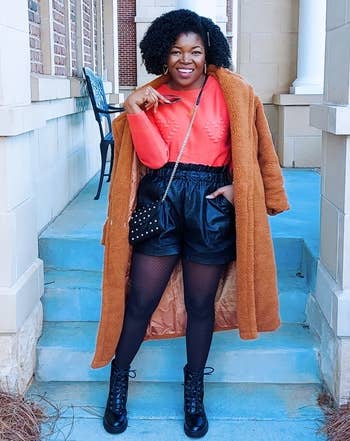 reviewer wearing the black shorts over tights with an orange top, coat, and boots