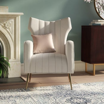 a white velvet wingback chair with a cream-colored throw pillow on it“class=
