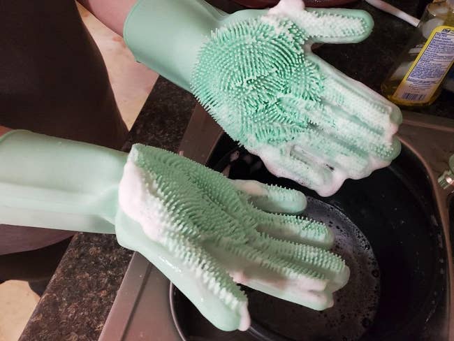 reviewer photo of them wearing the soapy, light teal gloves while washing dishes