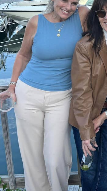 Two people standing by a dock. One wears a sleeveless top with wide pants; the other wears a brown jacket. Both hold drinks and smile at the camera
