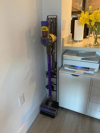 A cordless vacuum cleaner stands upright next to a printer on a floor in a home setting