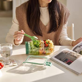 Woman eating crockery   from a jar with a greenish  fork astatine  a array  with a publication  and solid  of water, conveying a modern   casual eating  setting