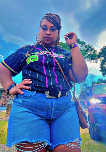 Person outdoors successful a casual outfit with a patterned shirt, denim shorts, belt, and a crossbody bag, striking a assured pose