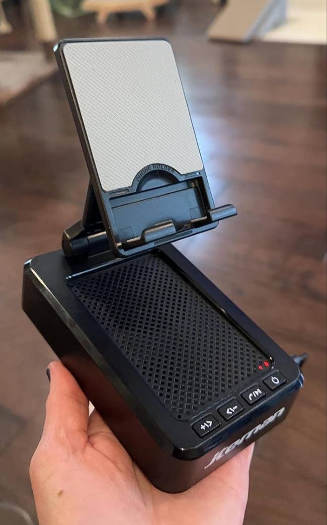 Hand holding a small black portable speaker with a stand for a phone attached to the top 