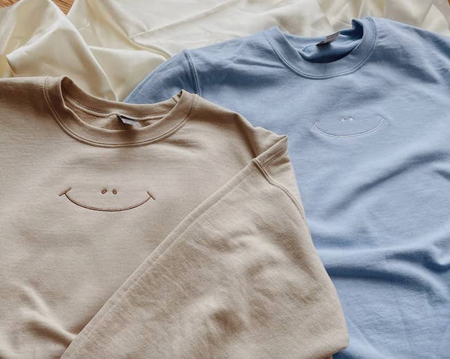 Two sweatshirts, one in a beige and another in blue, both have a small embroidered smiley face in the center