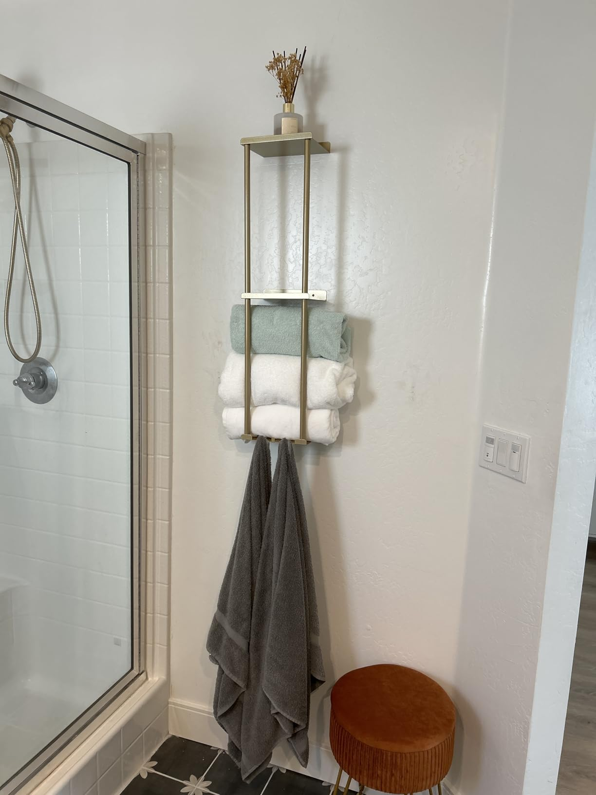 Wall-mounted bath  support  with neatly folded towels, a tiny  vase, and a grey  towel hanging below. A woody  stool is nearby