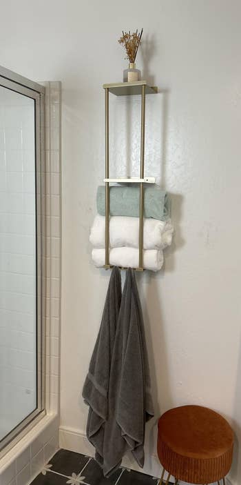 Wall-mounted bath support with neatly folded towels, a tiny vase, and a grey towel hanging below. A woody stool is nearby