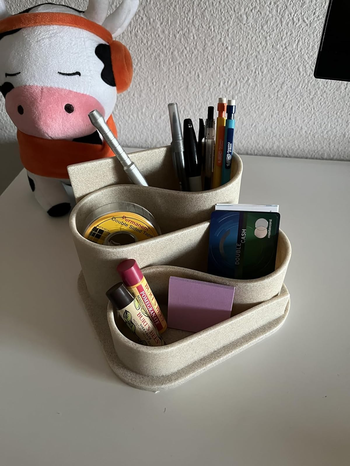 Desk organizer holding pens, CDs, a lipstick, sticky notes, and a wallet beside a plush cow toy on a table