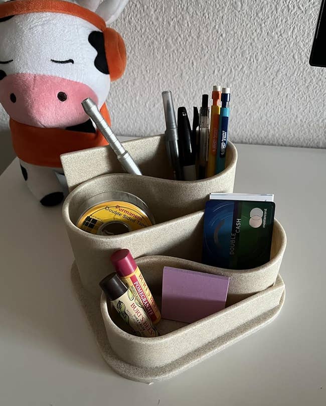 Desk organizer holding pens, CDs, a lipstick, sticky notes, and a wallet beside a plush cow toy on a table