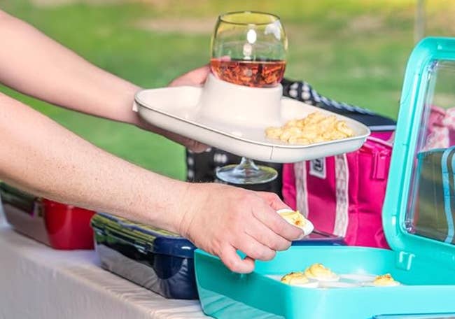 Person holding a tray with vino and snacks, reaching for nutrient from a cooler astatine an outdoor gathering