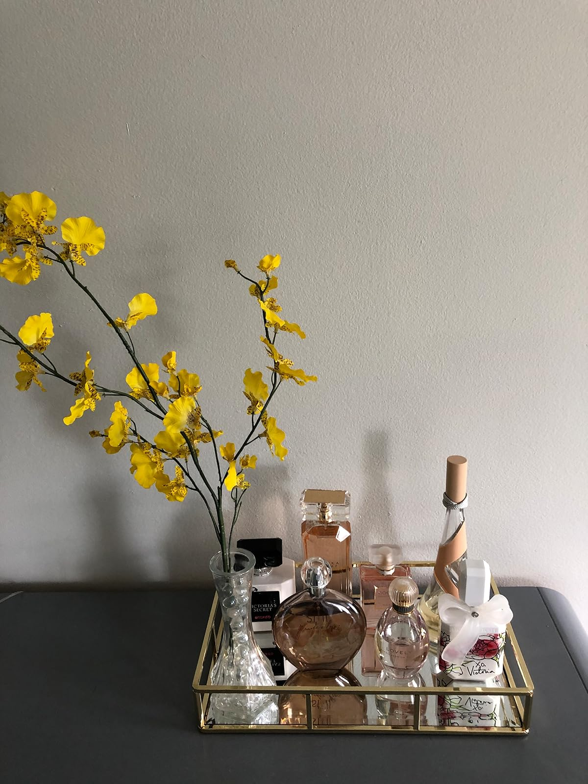 reviewer's mirrored tray with perfumes, a container  with a bow, and a vase with yellowish  flowers arranged neatly connected  a array  against a plain wall