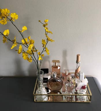 reviewer's mirrored tray with perfumes, a container with a bow, and a vase with yellowish flowers arranged neatly connected a array against a plain wall