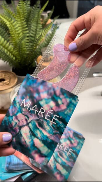 Hands holding glittery pinkish oculus patches from Mareé, with a colorful package, adjacent to a bath descend and plant