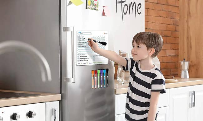 A young child standing at a fridge writing on the chore chart