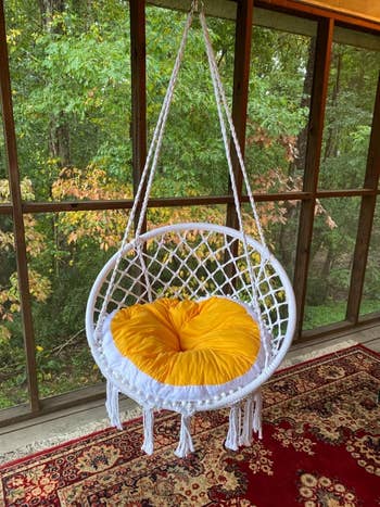 Hanging wicker chair with a yellow cushion on a porch, set against a forest view backdrop