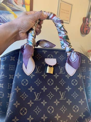 A hand holds a Louis Vuitton handbag with a patterned scarf wrapped around the handles