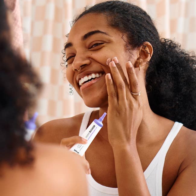 Woman applying skincare products, smiling at her reflection in a mirror