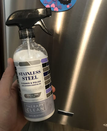 reviewer holding the bottle of the stainless steel cleaning spray with their stainless steel fridge in the background