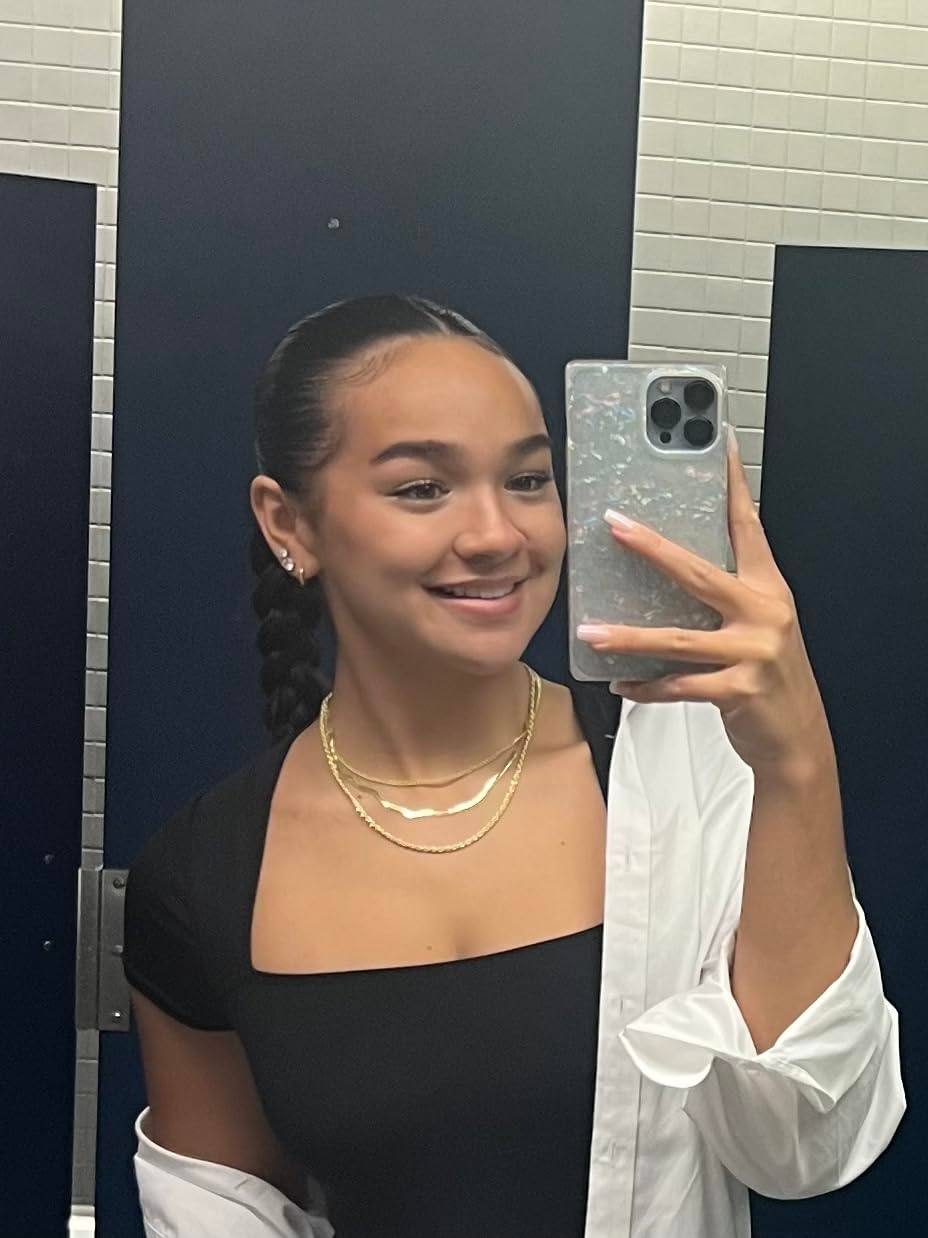 A woman takes a mirror selfie in a restroom, wearing a black top and layered necklaces, smiling as she holds a smartphone with a decorative case