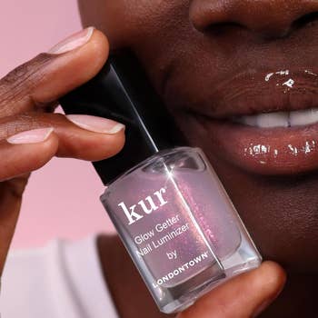 Person holding a vessel of kur Glow Getter Nail Luminizer by Londontown adjacent their lip, showcasing glossy nails and lips