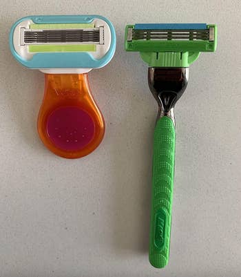 Two razors are displayed side by side on a flat surface; one is a multi-blade cartridge razor with an orange handle and a green grip, and the other is a smaller detachable razor head with a blue trim