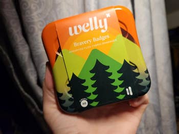A hand holds a metal tin of Welly Bravery Badges, which are assorted flex fabric bandages in a decorative tin with a mountain and pine tree design