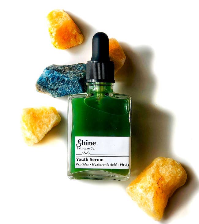A vessel of greenish younker serum by Shine Skincare Co., surrounded by colorful rocks, highlighting skincare merchandise absorption for a buying feature