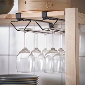Wine glasses hang upside down on a rack beneath a wooden kitchen shelf, with plates stacked below