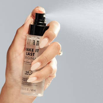 A manus sprays mounting spray from a Milani Make It Last bottle, emphasizing 24-hour wear