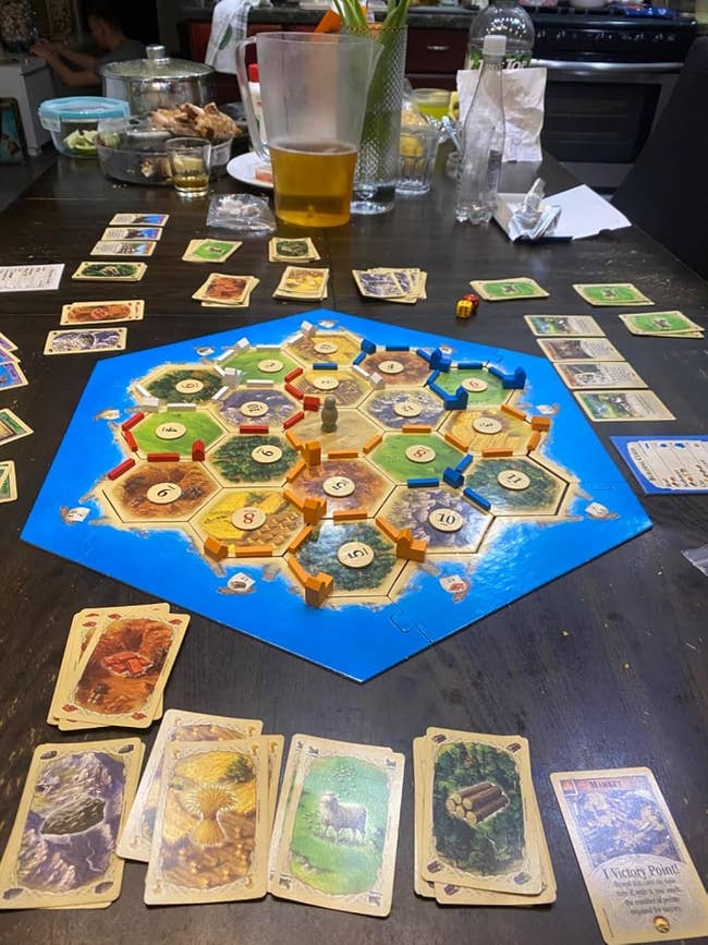 Board game Settlers of Catan in progress on a table, surrounded by cards, snacks, and drinks