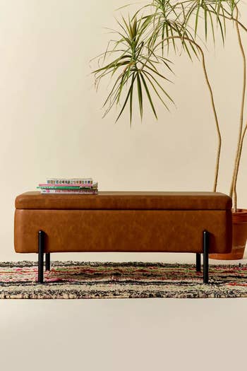 Tan leather bench with black legs on a patterned rug, next to a potted plant and a stack of books