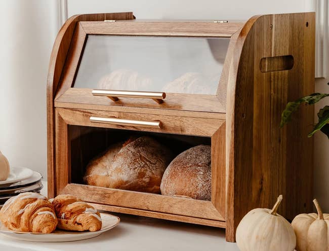 Wooden bread box with glass doors displaying loaves of bread and croissants, accompanied by stacked plates and decorative white pumpkins nearby