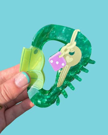 Hand holding a greenish hairsbreadth claw clip adorned with playful cardinal and dice embellishments, acceptable against a plain background