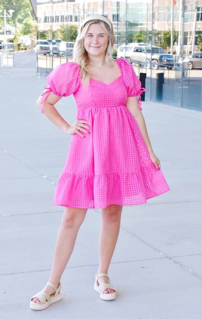 Model wearing hot pink puffy sleeved mini dress with skater skirt and grid pattern, paired with nude sandals outside