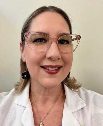 Person wearing glasses and a achromatic  garment  smiles astatine  the camera. They person  abbreviated  hairsbreadth  and are accessorized with earrings and a necklace