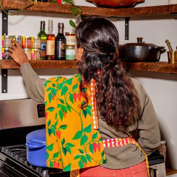 Person reaching for pantry items connected a room shelf, wearing a patterned apron with a plan featuring leaves and peppers