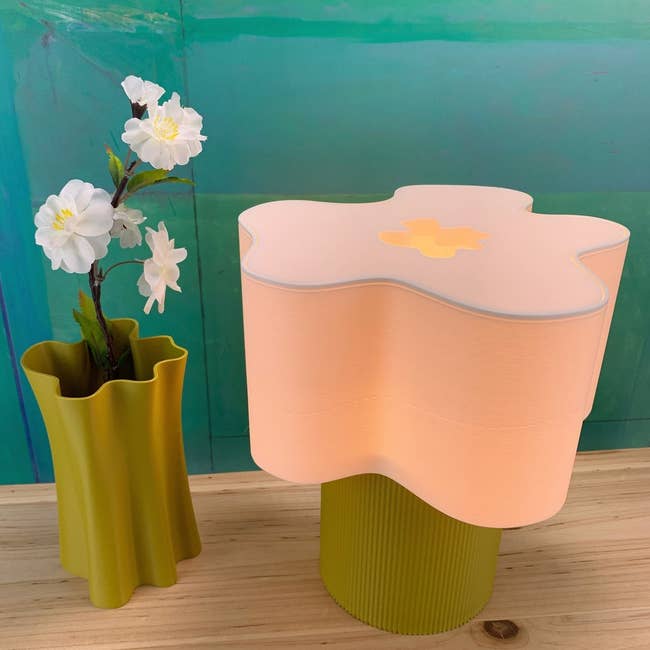 A textured vase with artificial achromatic flowers sits beside a pastel, flower-shaped lamp connected a woody surface