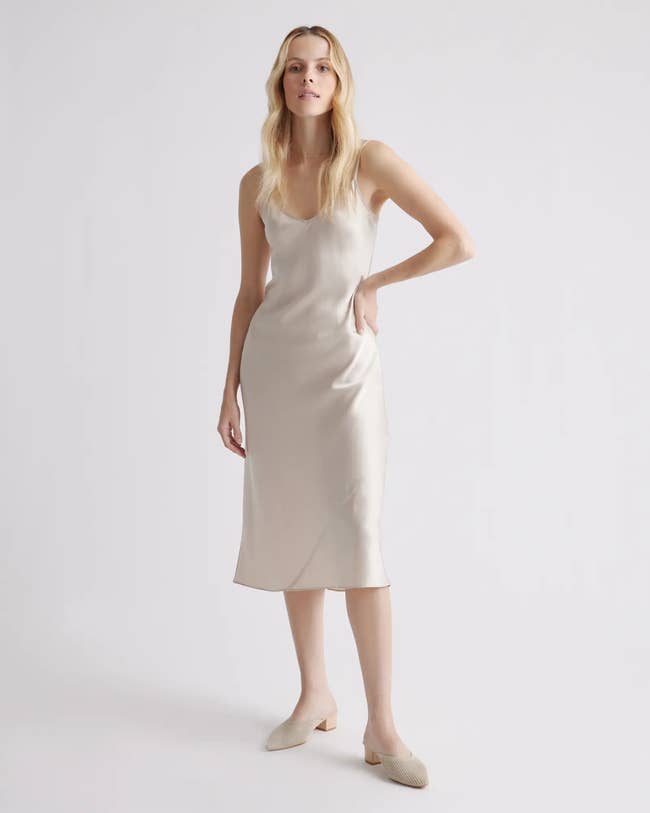 Model wearing midi-length white silk sleeveless dress with v-neckline and nude heeled mules on a white background