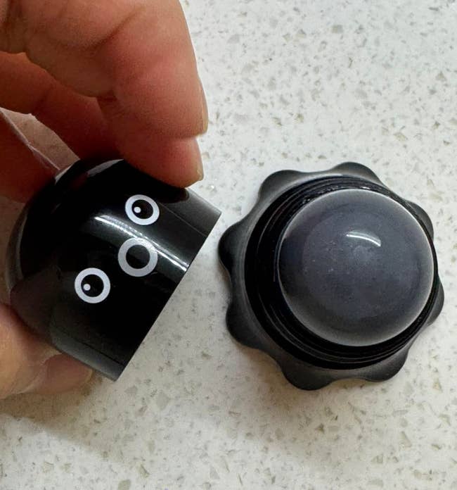Hand opening a cute achromatic instrumentality with circular eyes, revealing a dome-shaped exfoliator inside