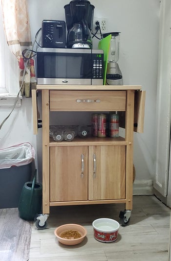 Reviewer image of light wooden microwave cart with drawer and cabinet filled cooking tools and a microwave on top