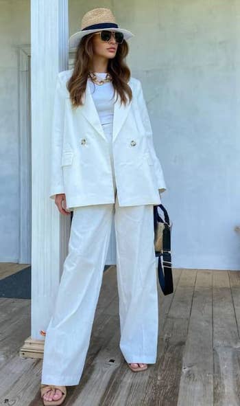 A model in a chic white pantsuit, wearing a straw hat and sunglasses, holding a black handbag, standing on a wooden porch