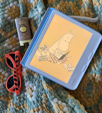 Kobo displaying a Moomin publication  sketch beside sunglasses and sunscreen, arranged connected  a patterned fabric. Suitable for summertime  buying  themes