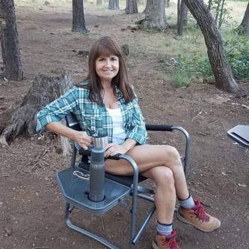 reviewer photo of them sitting in their black camper chair with the side table extended out and their water bottle in the built-in cupholder